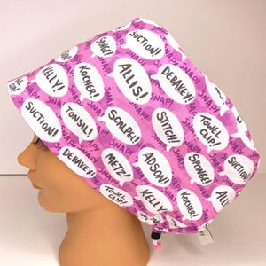 May include: A pink surgical scrub cap with white speech bubbles containing medical terms like "SUCTION!" and "SCALPEL!" The cap is designed to be worn in a medical setting and has a drawstring closure.