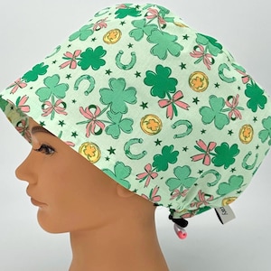 May include: A light green surgical scrub cap with a St. Patrick's Day theme. The cap features a repeating pattern of green shamrocks, pink bows, gold coins, horseshoes, and stars. The cap is tied at the back with a black drawstring.