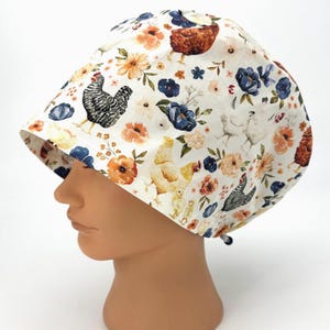 May include: A scrub cap featuring a cream-colored background with a repeating pattern of brown, white, and black chickens, chicks, and various orange, yellow, and blue flowers. The cap has a rounded crown and a tie closure at the back.
