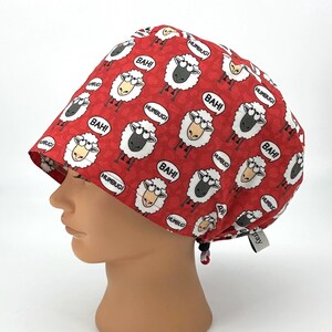 May include: Red surgical scrub cap with a repeating pattern of cartoon sheep. The sheep are white and black with speech bubbles that say "BAH!" and "HUMBUG!". The cap has a drawstring and a small tag that says "gray".
