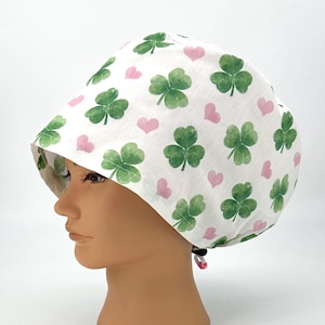 May include: White scrub cap with a pattern of green shamrocks and pink hearts. The cap is designed with a brim and a drawstring closure. The shamrocks are in a watercolor style, and the hearts are evenly spaced across the fabric.
