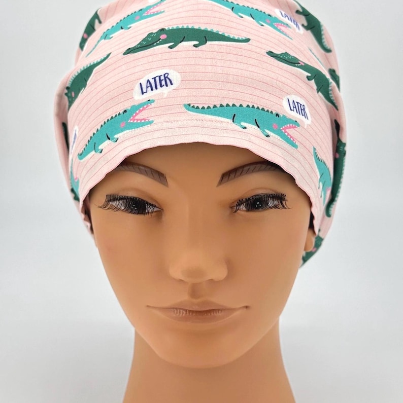 May include: A pink scrub cap with a pattern of green alligators and the word "LATER" in a speech bubble. The cap is made of a soft fabric and is designed to be worn in a medical setting.
