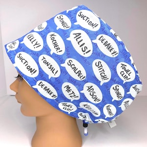 May include: A blue surgical scrub cap with white speech bubbles containing medical terms like "SUCTION!" and "SCALPEL!" The cap has an adjustable drawstring and is designed for medical professionals.
