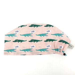 May include: A pink scrub cap with a repeating pattern of green alligators and the word "LATER" in speech bubbles. The cap has a striped background and a small gray tag.