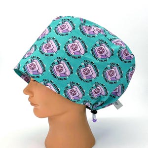 May include: A teal surgical scrub cap with a repeating pattern of purple rotary phones and the text "CALL ME ANTISOCIAL BUT PLEASE DON'T CALL ME." The cap has a drawstring and is designed to be worn during medical procedures.