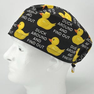 May include: Black scrub hat with a repeating pattern of yellow rubber ducks and the text "Duck Around and Find Out".