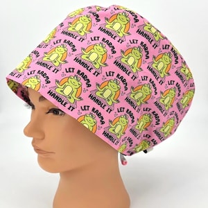 May include: Pink scrub cap with a repeating pattern of a meditating frog, the words "Let Karma Handle It", and small stars. The cap is designed to be worn in a medical or surgical setting.