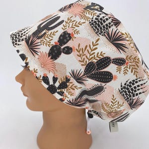May include: A white scrub cap featuring a desert cactus pattern in black, pink, and brown. The cap has a drawstring and a small tag. The design includes various cacti and floral elements.