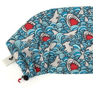 May include: A blue and white patterned scrub cap featuring a repeating design of gray sharks with open red mouths swimming in stylized blue waves. The cap has a drawstring and a small white tag with a black heart design.