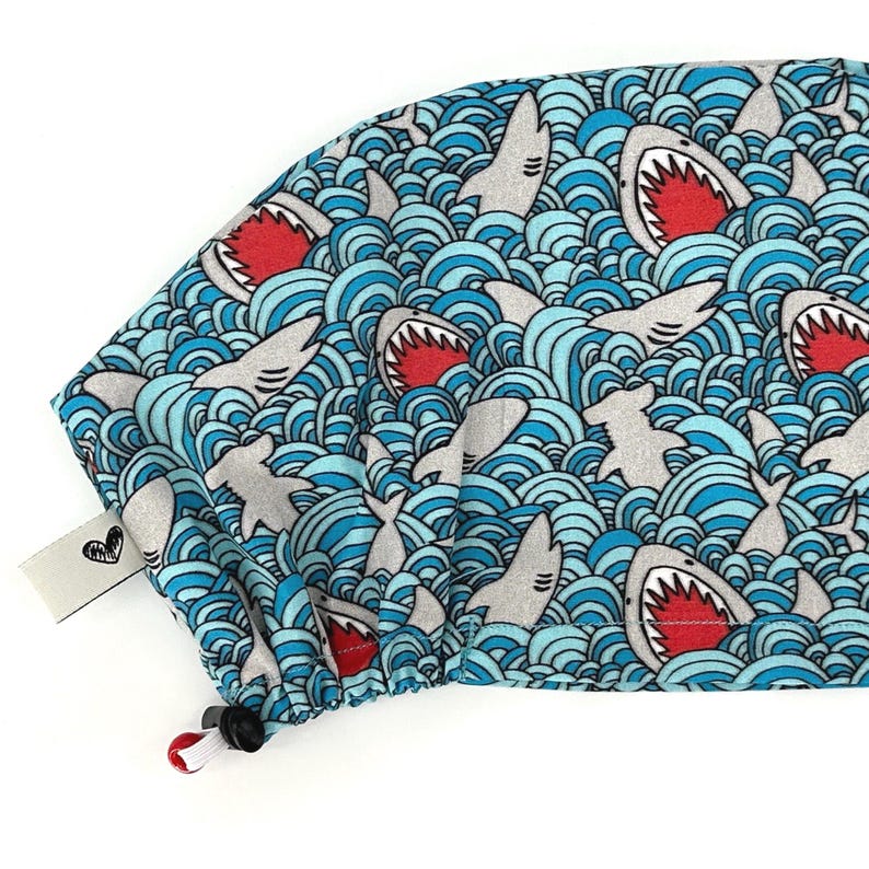 May include: A blue and white patterned scrub cap featuring a repeating design of gray sharks with open red mouths swimming in stylized blue waves. The cap has a drawstring and a small white tag with a black heart design.