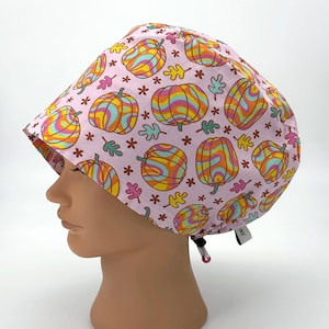 May include: A pink surgical scrub cap featuring a repeating pattern of colorful, psychedelic pumpkins, leaves, and stars. The pumpkins have orange, yellow, and blue swirls. The cap has a black drawstring and a small white tag.