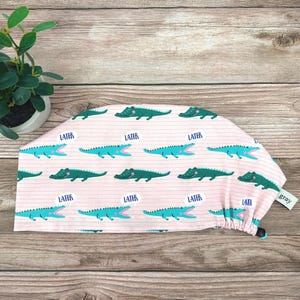 May include: A pink scrub cap with a repeating pattern of green alligators and the word "LATER" in blue speech bubbles. The cap has a drawstring and a small tag that says "gray".