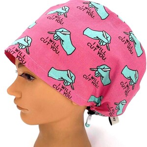 May include: A pink surgical scrub cap with a repeating pattern of teal-colored gloved hands holding scalpels, and the text "I WILL CUT YOU." The cap is tied at the back with an adjustable drawstring.