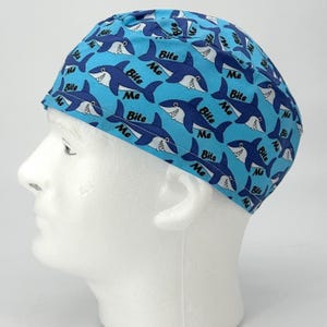 May include: A blue surgical scrub cap with a repeating pattern of cartoon sharks in navy blue and white. The text "Bite Me" is printed above each shark. The cap is made of a lightweight fabric.