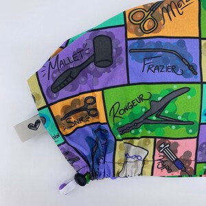 May include: A colorful fabric scrub cap with a variety of surgical tools and instruments printed on it. The cap has a drawstring and a black toggle. The text on the cap includes "Mallet", "Frazier", "Rongeur", "Snap", "Hypo", and "Metz".