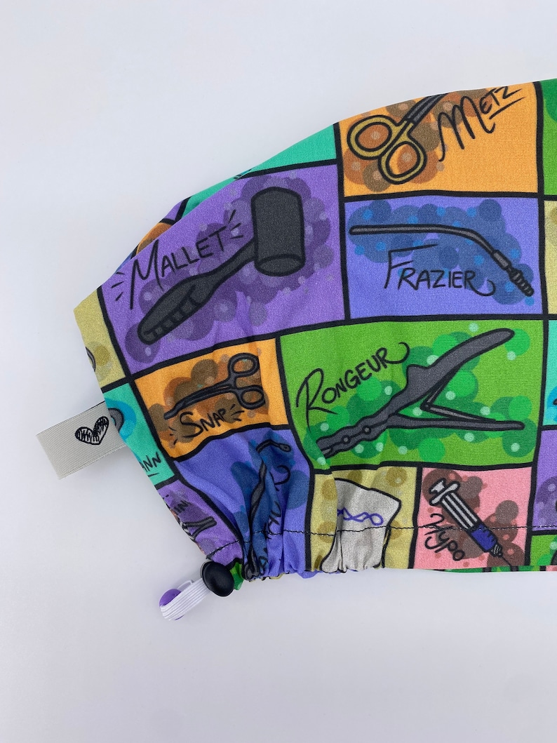 May include: A colorful fabric scrub cap with a variety of surgical tools and instruments printed on it. The cap has a drawstring and a black toggle. The text on the cap includes "Mallet", "Frazier", "Rongeur", "Snap", "Hypo", and "Metz".