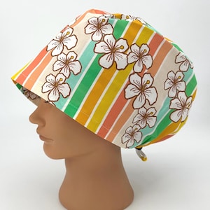 May include: A scrub cap featuring a pattern of white hibiscus flowers with brown outlines set against vertical stripes of orange, yellow, and green. The cap includes an adjustable toggle closure at the back to secure the fit around the head.