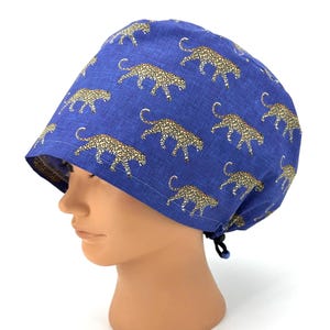May include: A blue surgical scrub cap featuring a repeating pattern of tan and black leopard print. The cap is made of fabric and has a black drawstring closure. The cap is shown on a mannequin head.
