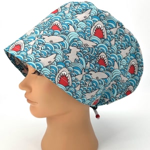 May include: A blue and white patterned hat featuring a repeating design of sharks and ocean waves. The sharks have red mouths and gray bodies. The hat is worn by a mannequin head.