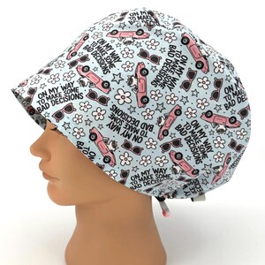 May include: A light blue surgical scrub cap with a repeating pattern of pink cars, sunglasses, white flowers, and the text "ON MY WAY TO MAKE SOME BAD DECISIONS". The cap has a curved brim and a tie in the back.