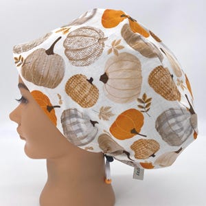 May include: A white scrub cap featuring a repeating pattern of pumpkins in shades of orange, brown, and tan. The pumpkins vary in style, with some appearing knitted or plaid. Autumn leaves are scattered among the pumpkins.