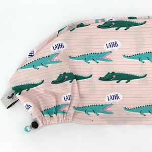 May include: A pink scrub cap with a repeating pattern of green alligators and the word "LATER" in speech bubbles. The cap has a drawstring and a small tag with a heart design.