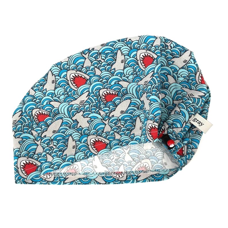 May include: A blue surgical scrub cap with a repeating pattern of gray sharks with red mouths swimming in blue waves. The cap has a tag that says "gray".