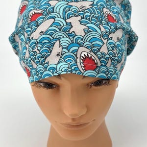 May include: A blue and white patterned scrub cap featuring a repeating design of sharks swimming in ocean waves. The sharks have gray bodies and red mouths. The cap is made of a soft, lightweight fabric.