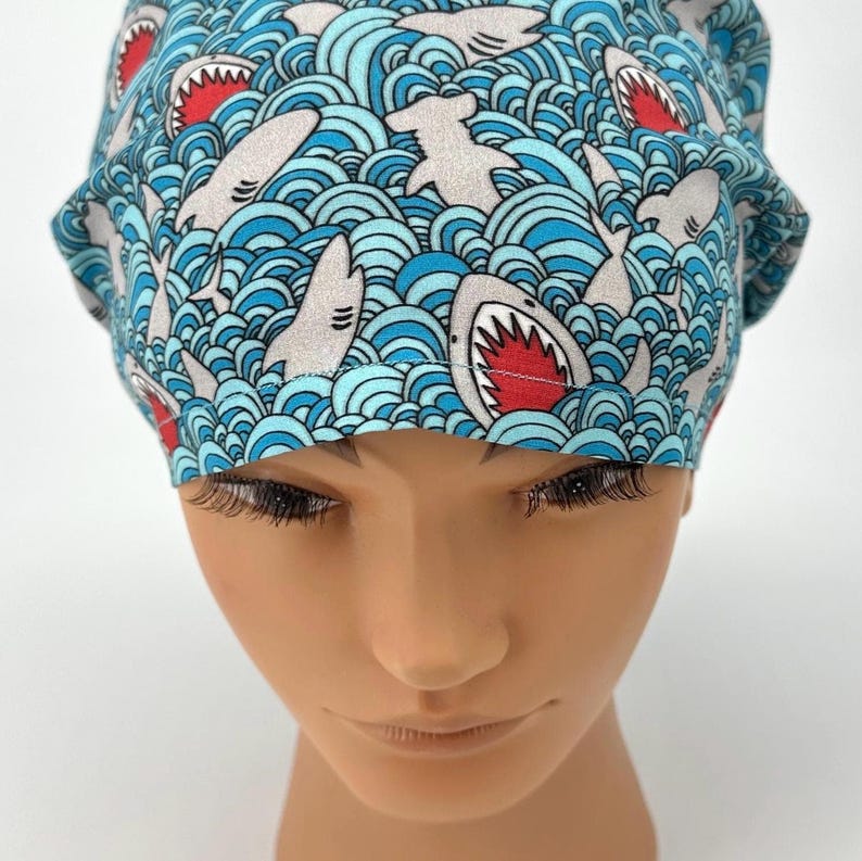 May include: A blue and white patterned scrub cap featuring a repeating design of sharks swimming in ocean waves. The sharks have gray bodies and red mouths. The cap is made of a soft, lightweight fabric.