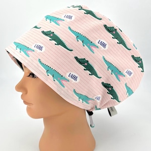 May include: A pink scrub cap with a repeating pattern of green and blue alligators and the word "LATER" in speech bubbles. The cap has a striped background and a tie closure.