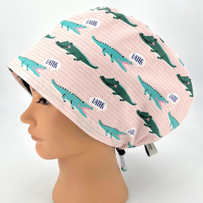 May include: A pink scrub cap with a repeating pattern of green and blue alligators and the word "LATER" in speech bubbles. The cap has a striped background and a tie closure.