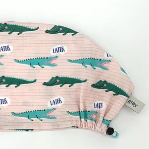 May include: A pink scrub cap with a repeating pattern of green alligators and speech bubbles that say "LATER". The cap has a drawstring and a small gray tag.