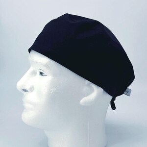 May include: A solid black surgical scrub cap, designed with a rounded crown and a tie closure at the back. The cap is displayed on a white mannequin head, showcasing its shape and fit. A small tag is attached to the cap.