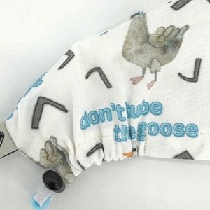 May include: White fabric with a blue goose and the text "don't tube the goose". The fabric has a black and white pattern of small arrows. The fabric is attached to a black button and a white and blue strap.