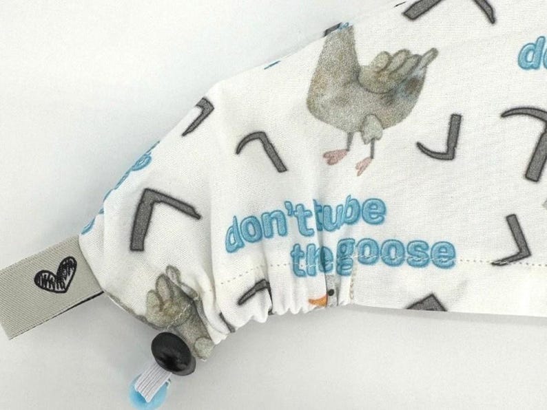 May include: White fabric with a blue goose and the text "don't tube the goose". The fabric has a black and white pattern of small arrows. The fabric is attached to a black button and a white and blue strap.
