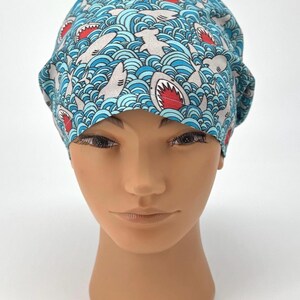May include: A blue surgical scrub cap with a repeating pattern of sharks swimming in wavy water. The sharks are gray with red mouths. The cap is made of a soft, lightweight fabric.