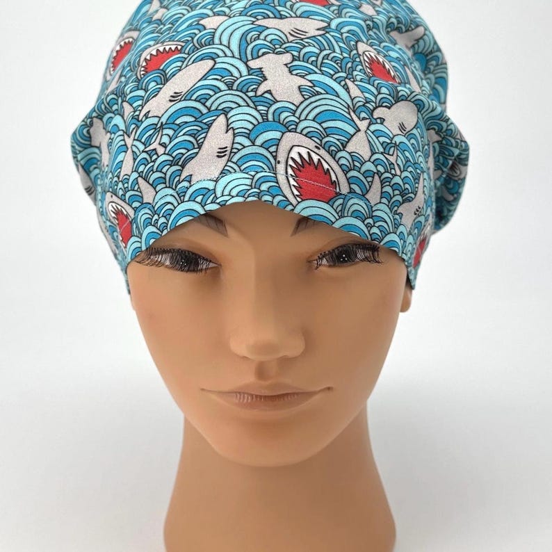 May include: A blue surgical scrub cap with a repeating pattern of sharks swimming in wavy water. The sharks are gray with red mouths. The cap is made of a soft, lightweight fabric.