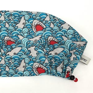 May include: A blue and white patterned surgical scrub cap featuring a repeating design of sharks in a wavy ocean. The sharks have open mouths with red interiors. The cap has a drawstring and a small gray tag.