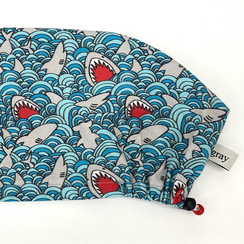 May include: A blue and white patterned surgical scrub cap featuring a repeating design of sharks in a wavy ocean. The sharks have open mouths with red interiors. The cap has a drawstring and a small gray tag.