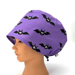 May include: A purple surgical scrub cap featuring a repeating pattern of black cartoon bats. The cap is tied at the back and is made of a lightweight fabric. The design is suitable for Halloween or a medical setting.