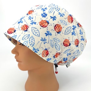 May include: A white surgical scrub cap featuring a repeating pattern of red ladybugs with white spots, blue leaves, and blue four-leaf clovers. The cap is tied at the back with a black and white string.