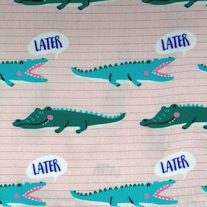 May include: Pink fabric with a repeating pattern of cartoon alligators in teal and green. Each alligator has an open mouth and a pink tongue. White speech bubbles with the word "LATER" are scattered throughout the design.