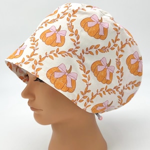 May include: A scrub cap featuring an off-white background with a repeating pattern of orange pumpkins adorned with light pink bows, connected by thin orange leafy vines. The cap has a rounded crown and an adjustable toggle closure at the back.