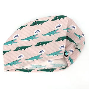 May include: A pink scrub cap with a repeating pattern of green and teal alligators and speech bubbles that say "LATER". The cap has a button closure and a small gray tag.