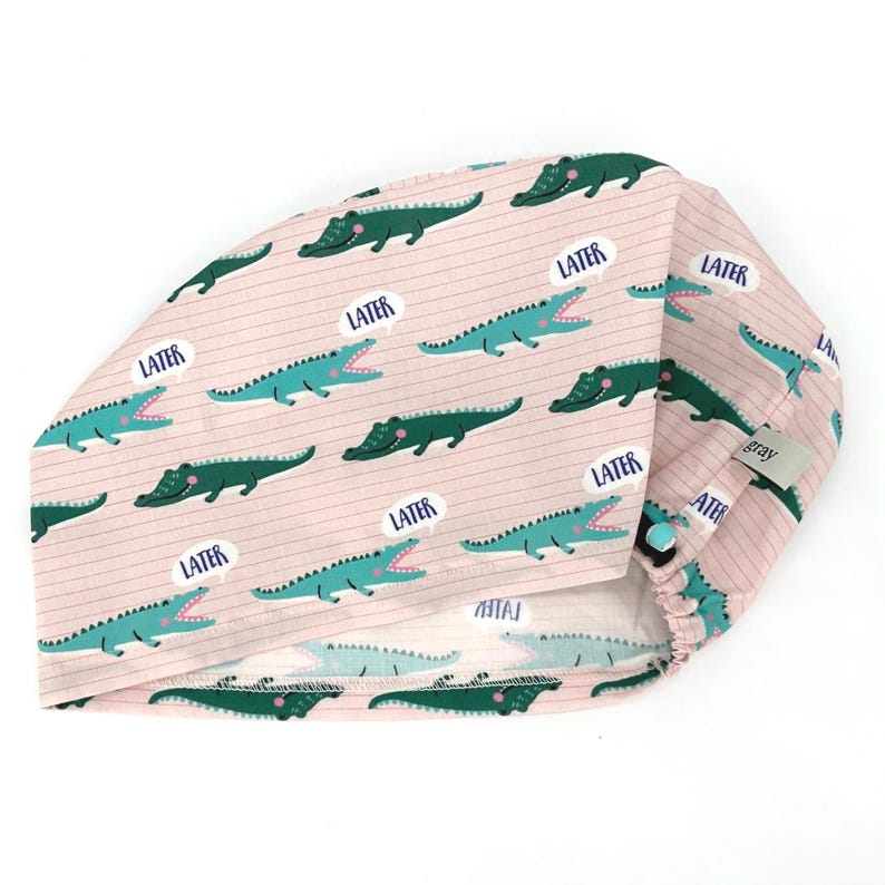 May include: A pink scrub cap with a repeating pattern of green and teal alligators and speech bubbles that say "LATER". The cap has a button closure and a small gray tag.