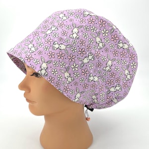 May include: A light purple scrub cap featuring a repeating pattern of white bunnies and pink and white flowers. The cap is tied in the back with a black drawstring and is designed to cover the hair.