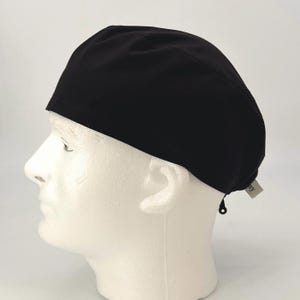 May include: A black surgical scrub cap made of a soft, opaque fabric. The cap is shown on a white mannequin head. A small tag is attached to the cap's edge. The cap is designed to cover the head and secure hair during medical procedures.