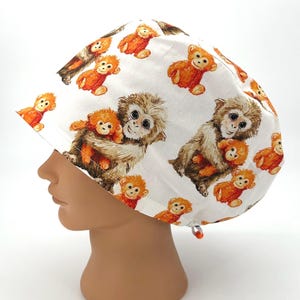 May include: White surgical scrub cap featuring a repeating pattern of brown and orange monkeys. The cap has a drawstring closure and is designed to cover the head. The monkeys are depicted in a cartoon style.