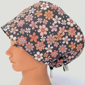 May include: A scrub cap featuring a dark gray background with a repeating pattern of five-petaled flowers in shades of orange, light pink, and white, accented by small white stars. The cap includes an adjustable toggle closure with a small gray tag.