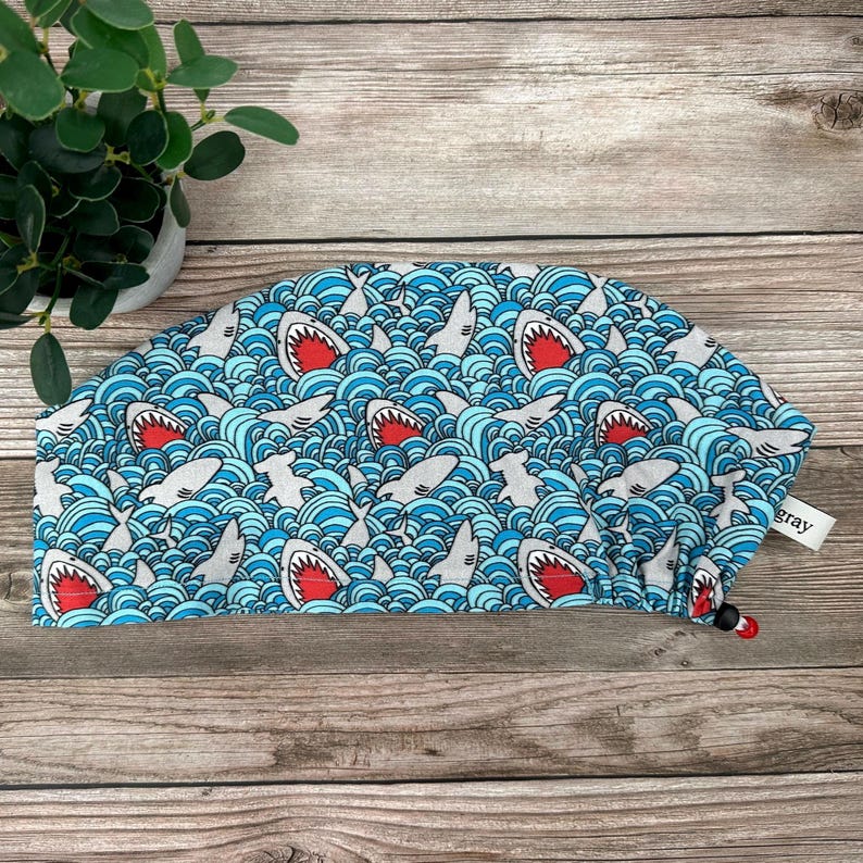 May include: A blue and white patterned surgical scrub cap featuring a repeating design of cartoon sharks with red mouths swimming in wavy water. The cap has a drawstring and a small tag that says "gray".